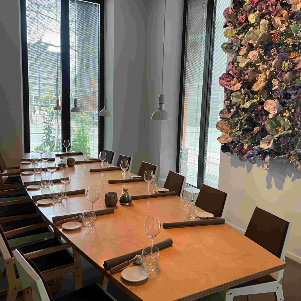 Strakke eetruimte met lange houten tafel, gedekt met glazen en rolplacemats, en een kleurrijk 3D bloemenkunstwerk.