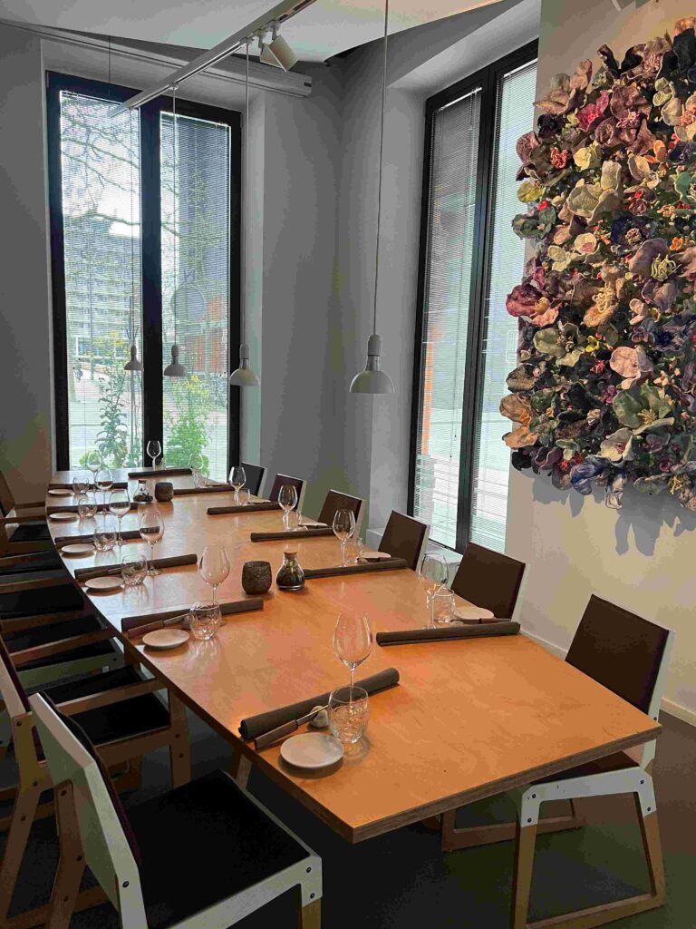 Strakke eetruimte met lange houten tafel, gedekt met glazen en rolplacemats, en een kleurrijk 3D bloemenkunstwerk.