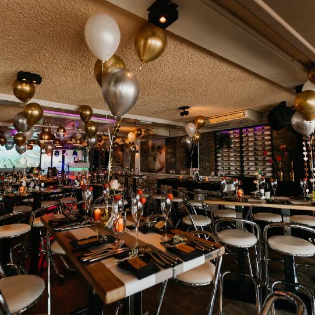 Sfeervolle evenementenlocatie met goud- en zilverkleurige ballonnen, gedekte tafels en barkrukken.