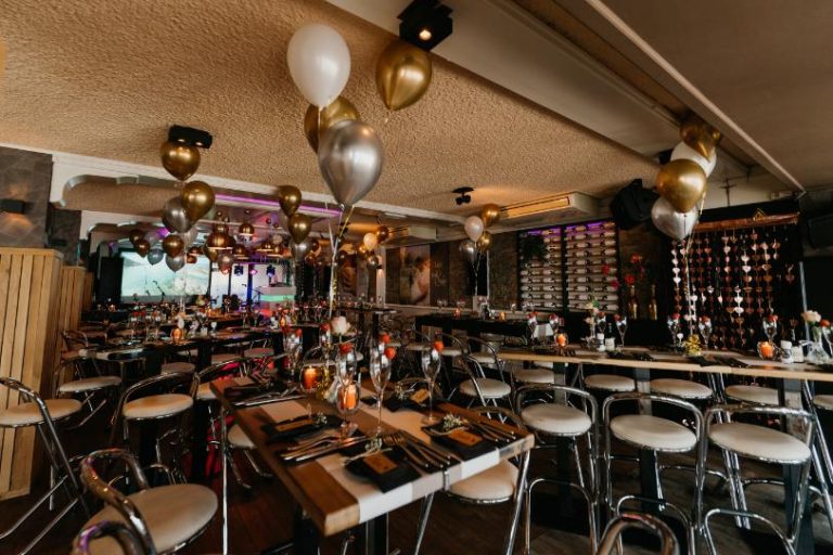Sfeervolle evenementenlocatie met goud- en zilverkleurige ballonnen, gedekte tafels en barkrukken.