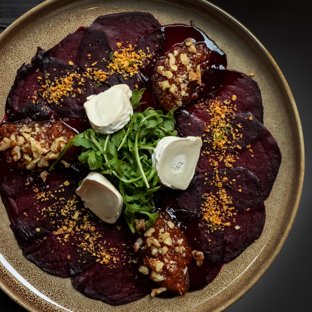 Bietencarpaccio met geitenkaas, vijgencompote, walnoten, rucola en goudgele kruimels op een beige bord.