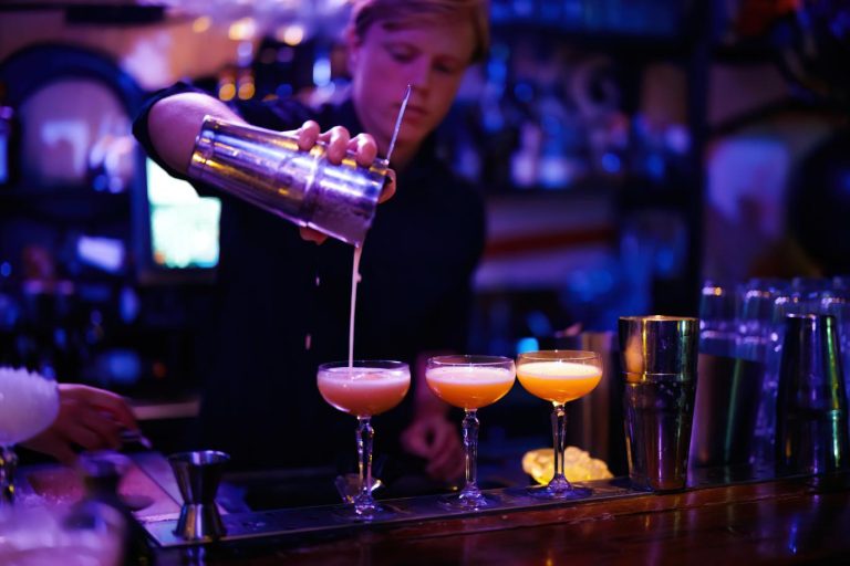 Blonde barman schenkt een oranje cocktail uit een shaker in een coupe glas op een sfeervolle bar.