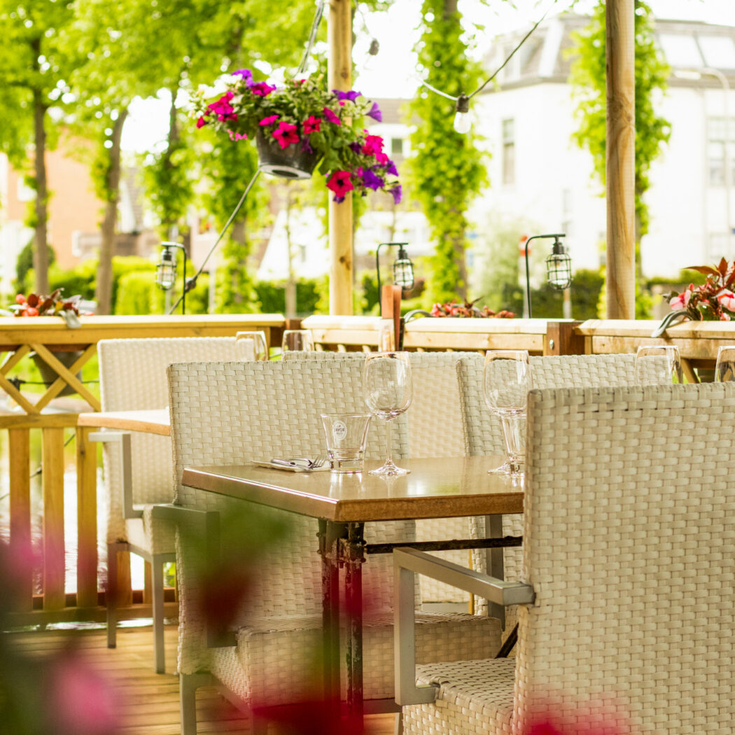 Sfeervol restaurantterras met gedekte tafels, gevlochten stoelen, een hangende bloempot en SWINCKELS parasol.