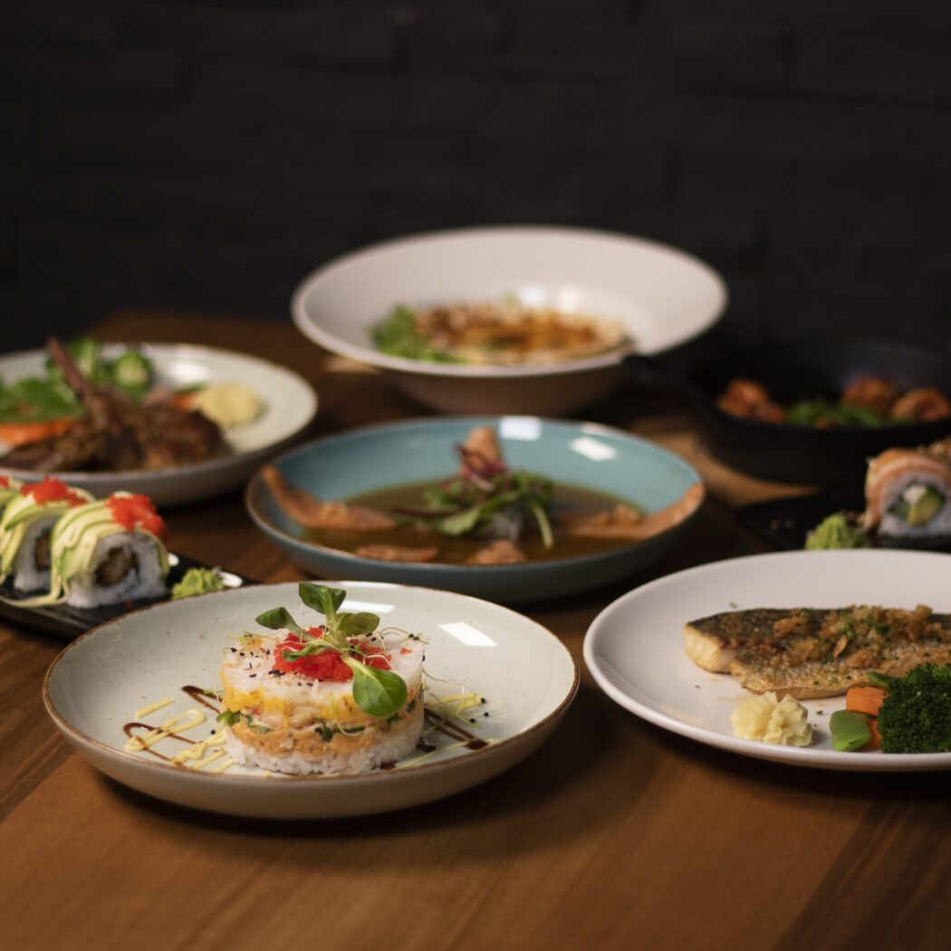 Een houten tafel vol met diverse restaurantgerechten: sushi, gegrilde vis, lamskoteletten en een rijsttorentje.