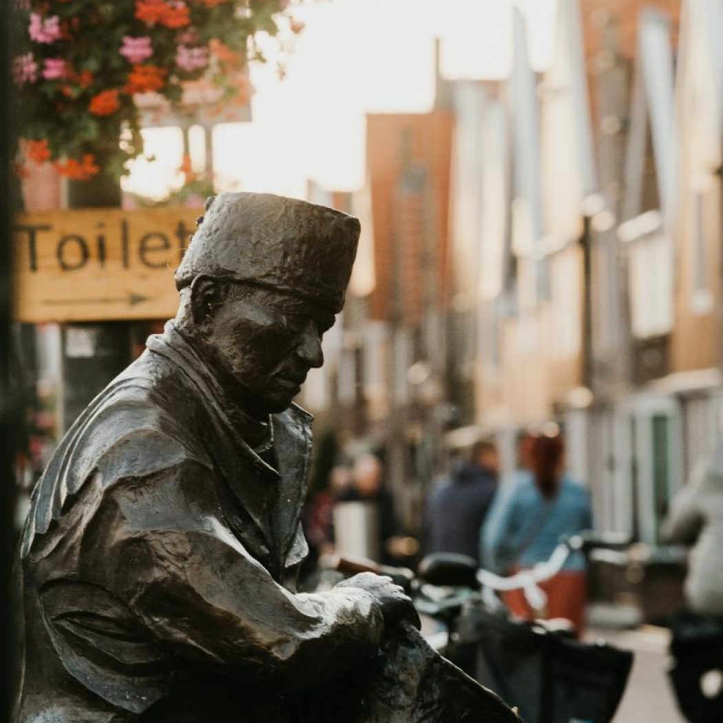 Bronzen beeld van zittende man met fez en tas op straat, bij Toilet-bord en overhangende roze-rode bloemen.