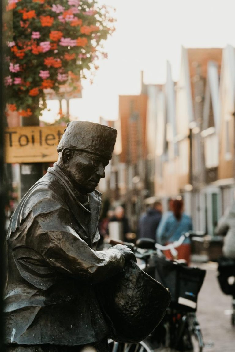 Bronzen beeld van zittende man met fez en tas op straat, bij Toilet-bord en overhangende roze-rode bloemen.