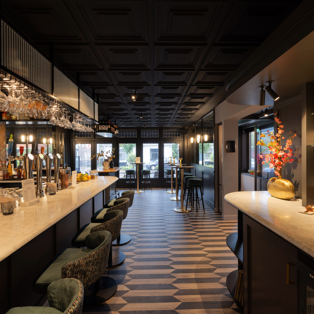Modern barinterieur met lange marmeren toonbank, groene barkrukken, hangende glazen en BITES EN DRINKS menu op een geometrische vloer.