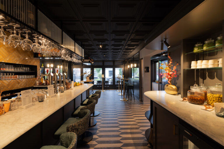 Modern barinterieur met lange marmeren toonbank, groene barkrukken, hangende glazen en BITES EN DRINKS menu op een geometrische vloer.
