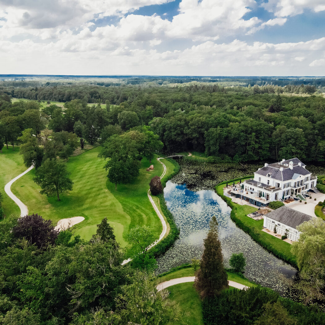 Luchtfoto van een elegant wit landhuis aan het water, geflankeerd door een groene golfbaan en dicht bos.