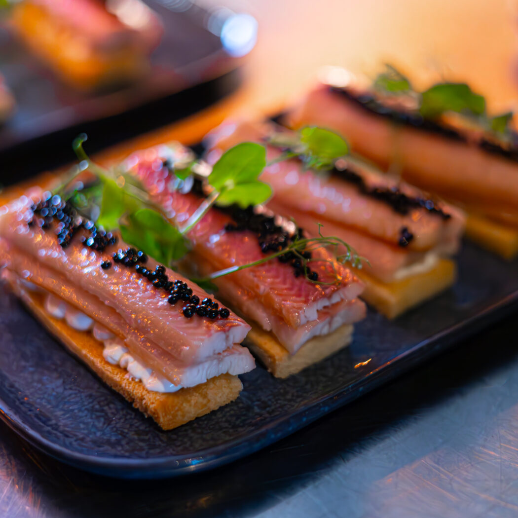 Vier luxe hapjes met gerookte vis, zwarte kaviaar en microgroenten op een donkerblauw bord.