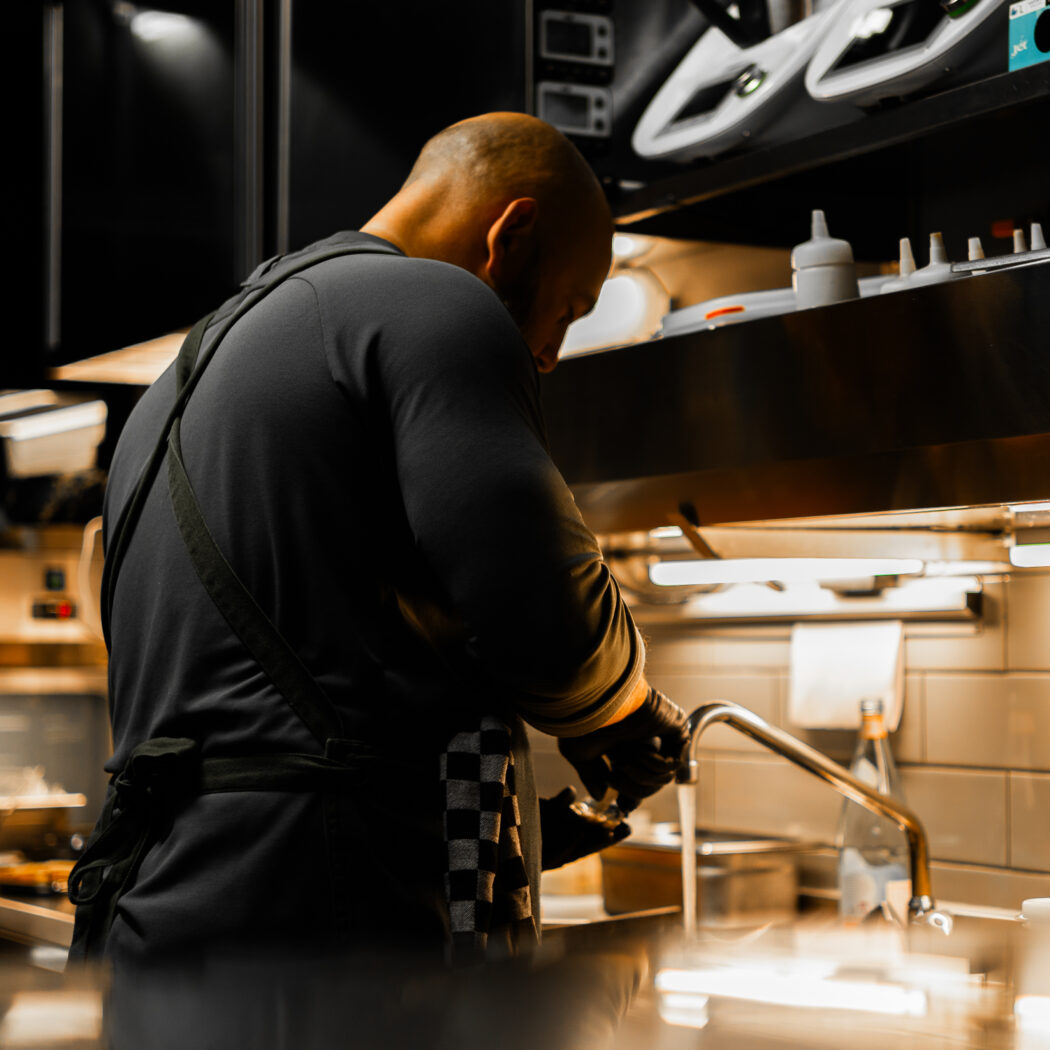 Kale chef met zwarte handschoenen wast onder de kraan in een professionele keuken met witte tegels.