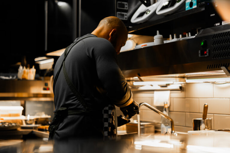 Kale chef met zwarte handschoenen wast onder de kraan in een professionele keuken met witte tegels.
