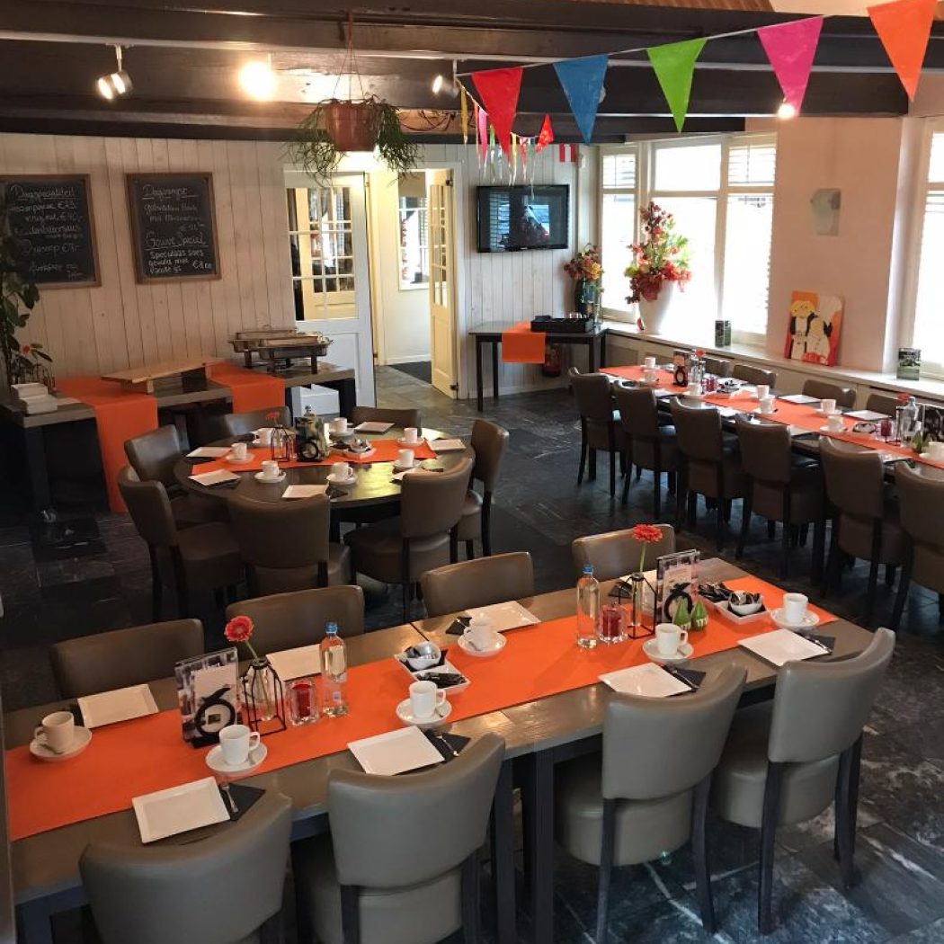 Gezellig ingerichte feestruimte met gedekte tafels, oranje lopers en kleurrijke vlaggetjes.