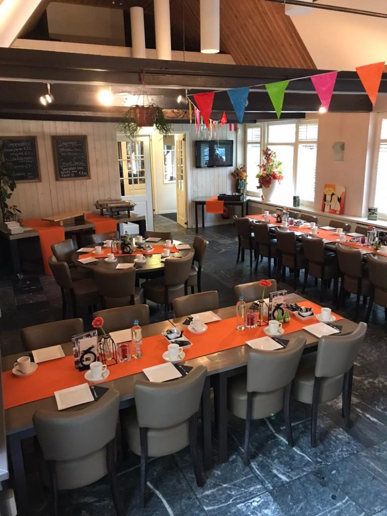 Gezellig ingerichte feestruimte met gedekte tafels, oranje lopers en kleurrijke vlaggetjes.