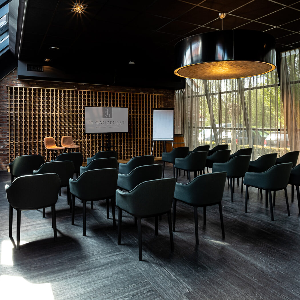 Moderne conferentiezaal met rijen donkergroene stoelen, scherm met T GANZENNEST logo en flipover.