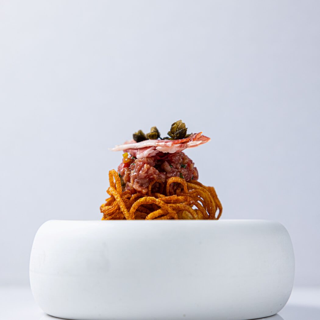 Rundertartaar op krokante gouden frietslierten, gegarneerd met ham en kappertjes in een moderne witte kom.