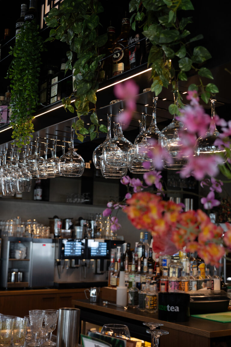 Barinterieur met hangende glazen, koffiehoek, diverse drankflessen, groene planten, roze bloemen en FUZETEA op toonbank.