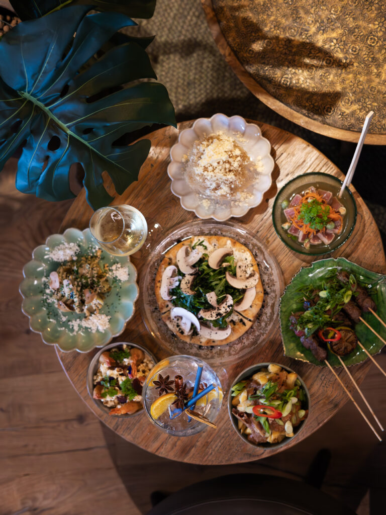Overzicht van een houten tafel met diverse kleine gerechten, waaronder spiesjes, flatbread, salades, een cocktail en witte wijn.