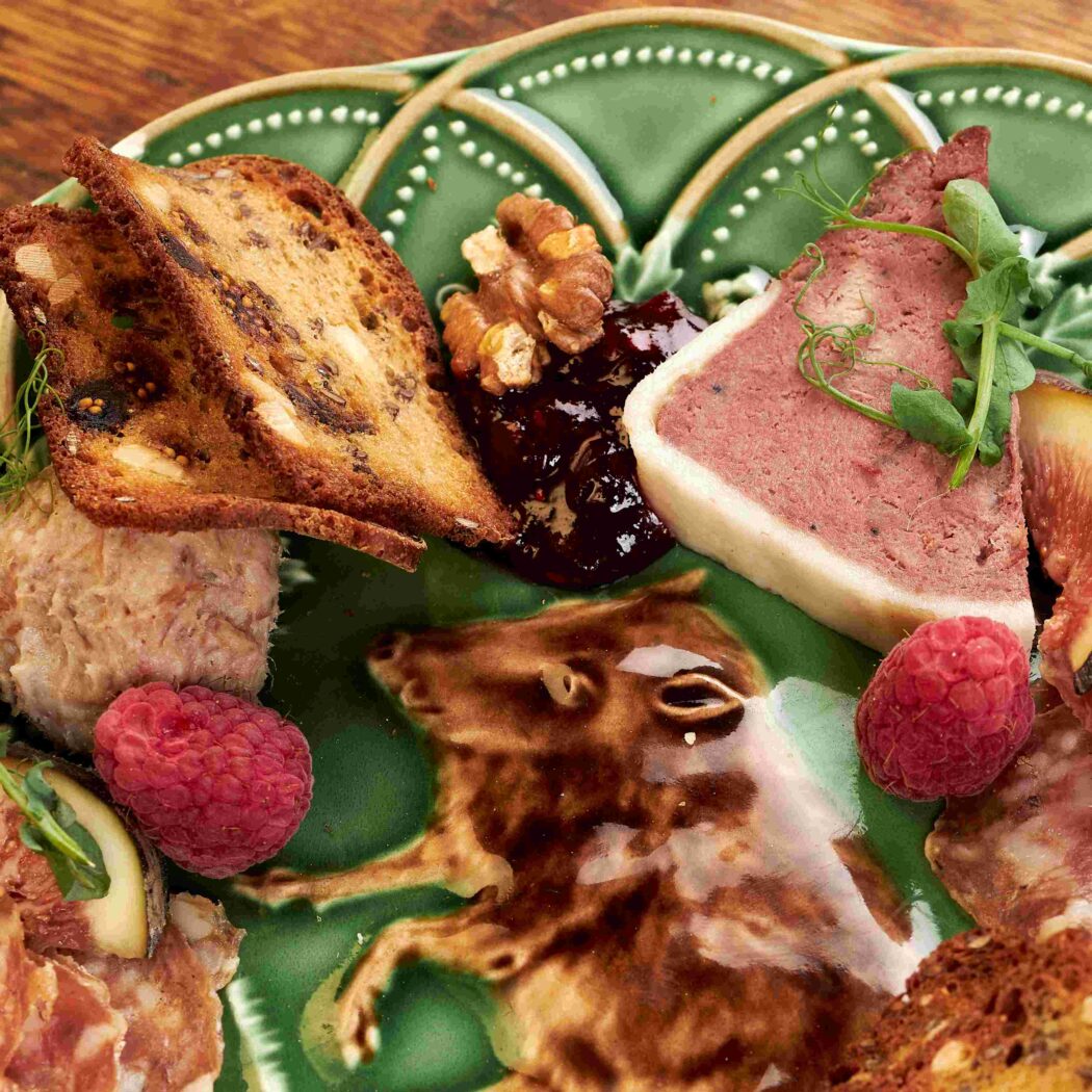 Charcuterieplank met terrine, toast, diverse vleeswaren, jam, frambozen, walnoten en vijgen op een groene schaal met varkensreliëf.