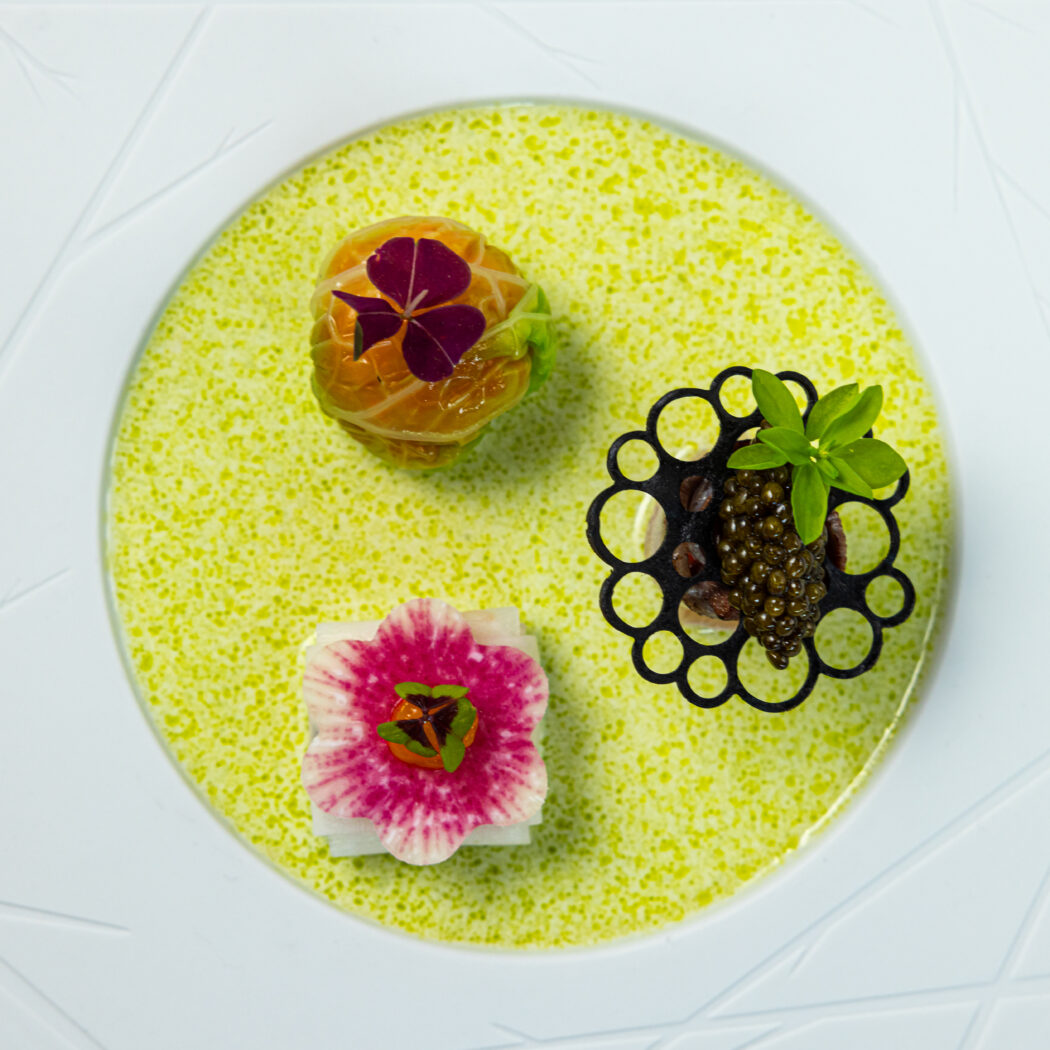 Drie kunstzinnige amuses op wit bord met groene saus: kaviaar, een geeloranje bol met paarse bloem, en een roze-witte bloem.