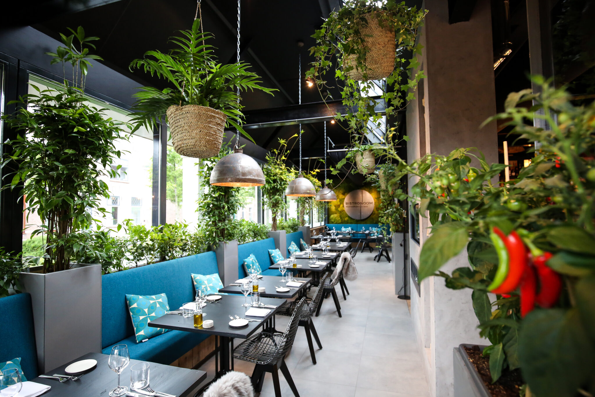 Stijlvol restaurantinterieur met turquoise banken, donkere tafels, weelderige planten, industriële lampen en BISTRONOOM logo.