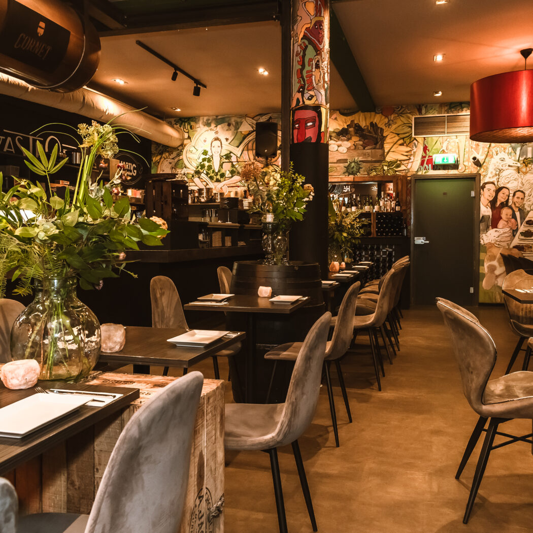 Sfeervol restaurant met donkere tafels, fluwelen stoelen, planten, kleurrijke muurschilderingen, een CORNET vat en een GASTRO bord.