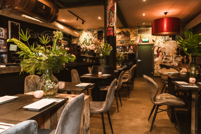 Sfeervol restaurant met donkere tafels, fluwelen stoelen, planten, kleurrijke muurschilderingen, een CORNET vat en een GASTRO bord.