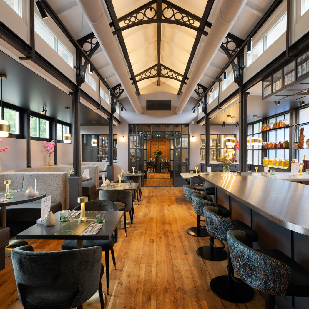 Licht restaurantinterieur met houten vloer, moderne bar, zitbanken en eettafels, onder een hoog plafond met spanten.