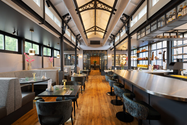 Licht restaurantinterieur met houten vloer, moderne bar, zitbanken en eettafels, onder een hoog plafond met spanten.