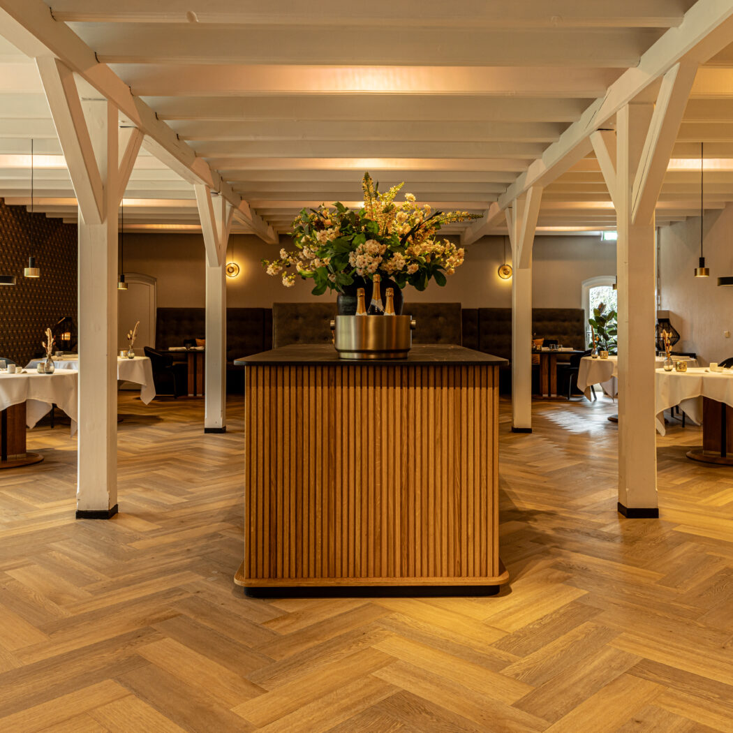 Elegant restaurantinterieur met houten visgraatvloer, witte balken en een centrale balie met bloemstuk en champagne.