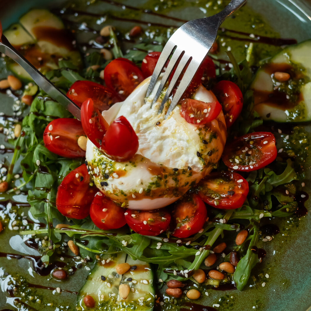 Hand snijdt romige burrata met mes en vork in een salade met cherrytomaten, rucola, pijnboompitten en pesto op groene schaal.