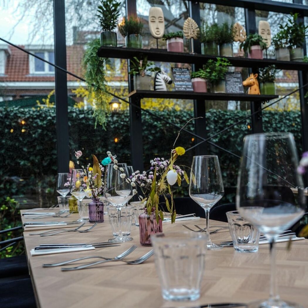 Gedekte houten tafel met visgraatmotief, glazen, bestek en lentebloemen in een restaurant met plantenplanken en grote ramen.