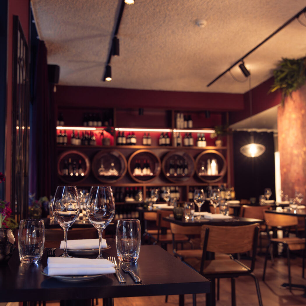 Sfeervol restaurantinterieur met gedekte tafels, wijnglazen en flessen wijn in schappen en houten tonnen.