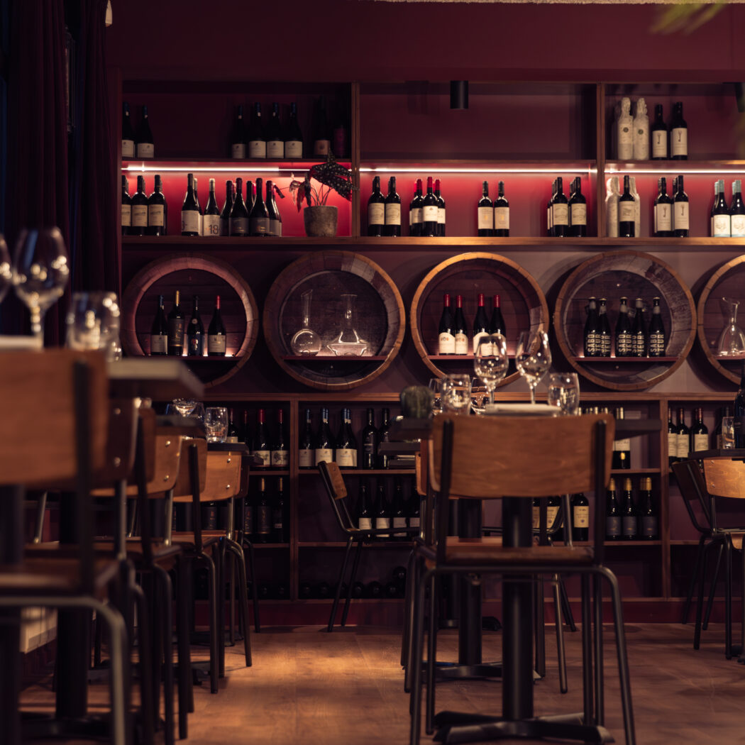 Sfeervolle donkerrode wijnbar met houten tafels en een achterwand vol verlichte wijnflessen en karaffen.