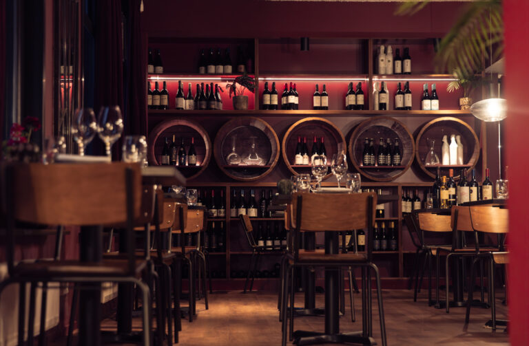 Sfeervolle donkerrode wijnbar met houten tafels en een achterwand vol verlichte wijnflessen en karaffen.