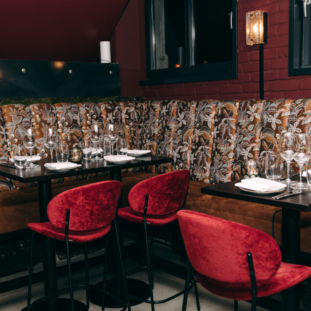 Sfeervol restaurant interieur met gedekte tafels, een bruine bank met bladpatroon en rode fluwelen barkrukken.