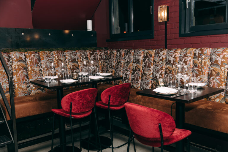 Sfeervol restaurant interieur met gedekte tafels, een bruine bank met bladpatroon en rode fluwelen barkrukken.