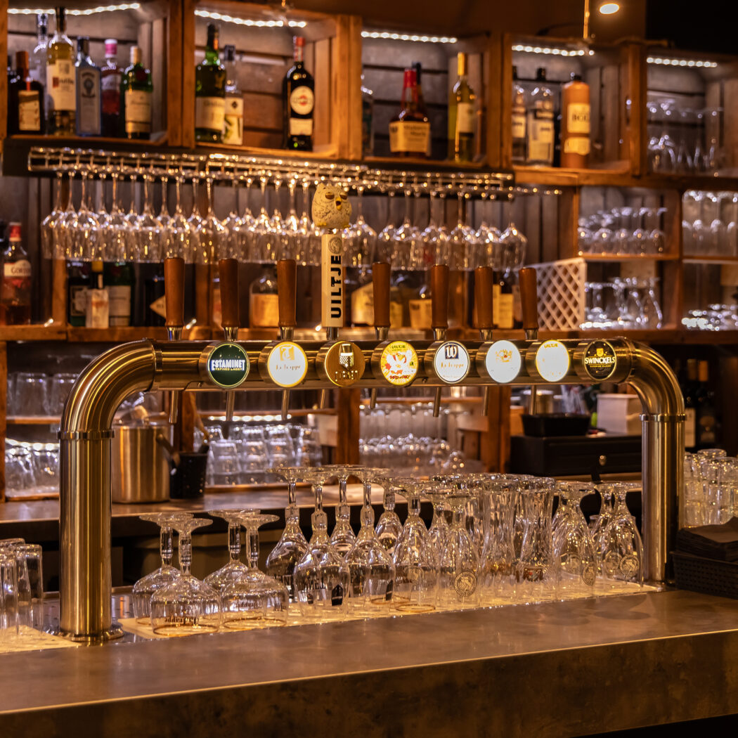 Glinsterende bar met biertappen zoals UILTJE en La Trappe, glazen, en houten rekken met verlichte drankflessen.