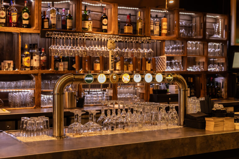 Glinsterende bar met biertappen zoals UILTJE en La Trappe, glazen, en houten rekken met verlichte drankflessen.