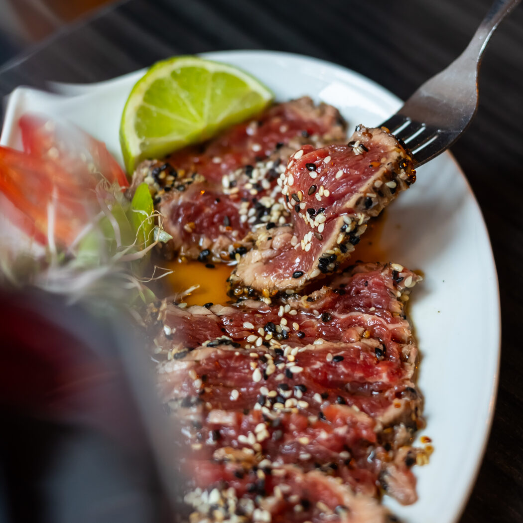 Vork tilt een stukje dun gesneden tonijn tataki met sesamzaadjes op van een wit bord met limoen.