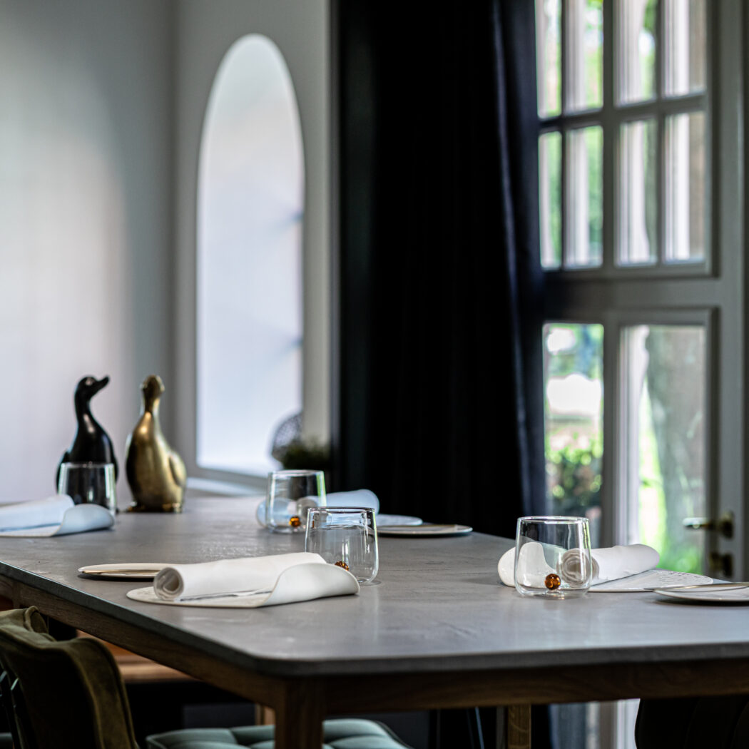 Elegante donkergrijze eettafel gedekt met witte servetten, glazen en twee decoratieve eenden, verlicht door grote ramen.