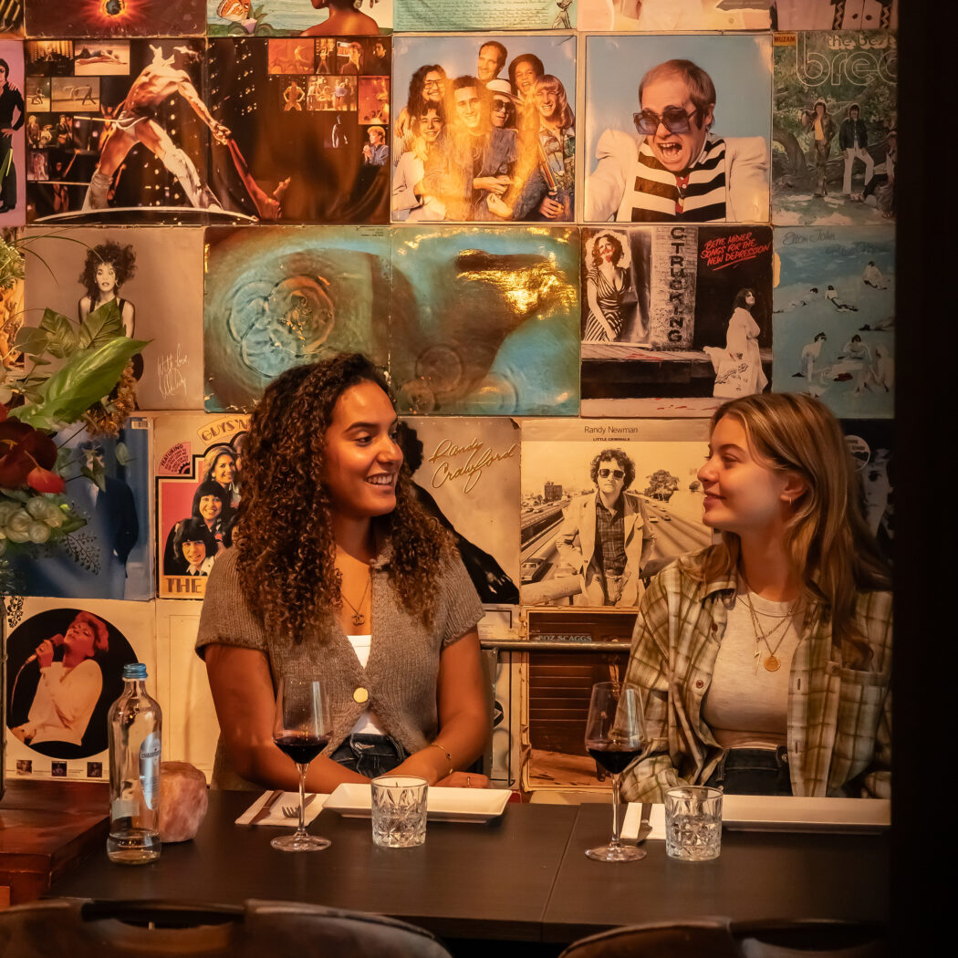 Twee lachende vrouwen zitten aan tafel in een restaurant met een wand van vinylhoezen, glazen rode wijn voor zich.