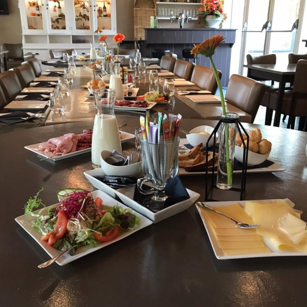 Lange tafel in een restaurant, gedekt met ontbijtgerechten, kannen melk en oranje gerberabloemen.