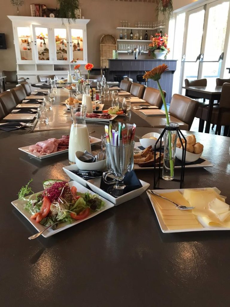 Lange tafel in een restaurant, gedekt met ontbijtgerechten, kannen melk en oranje gerberabloemen.