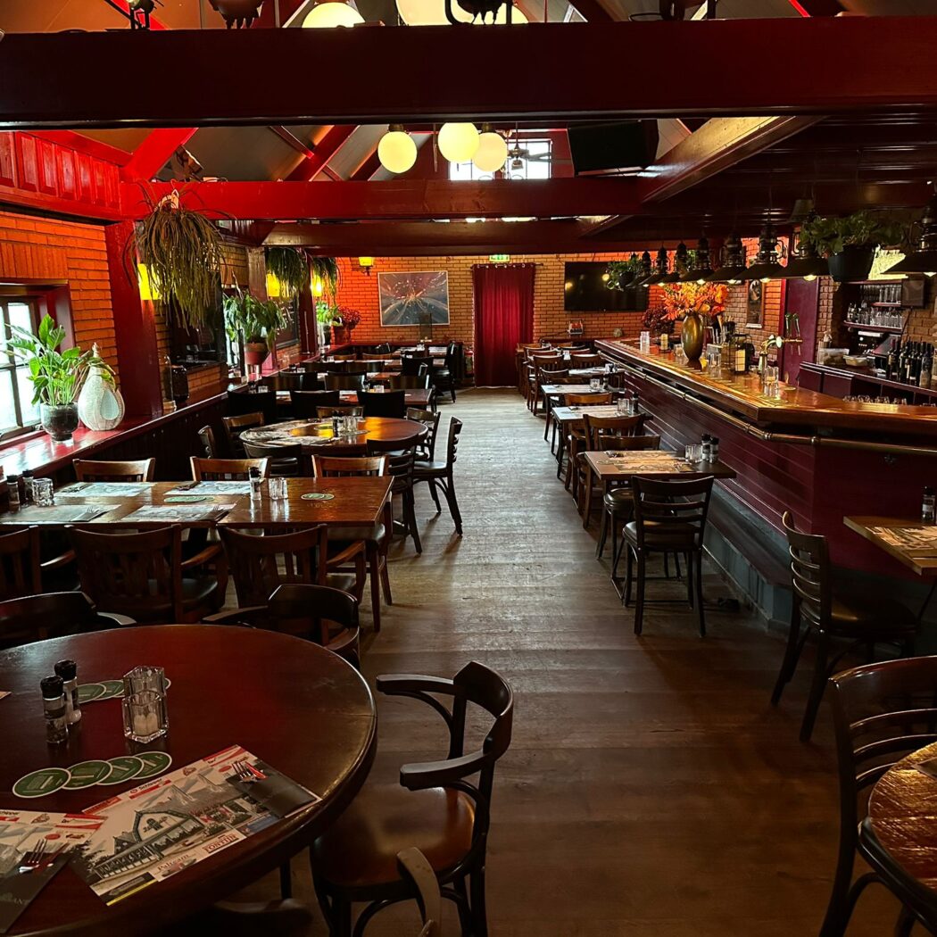 Sfeervol restaurant met donkere houten tafels, rode bakstenen muren, een lange bar en hangplanten.