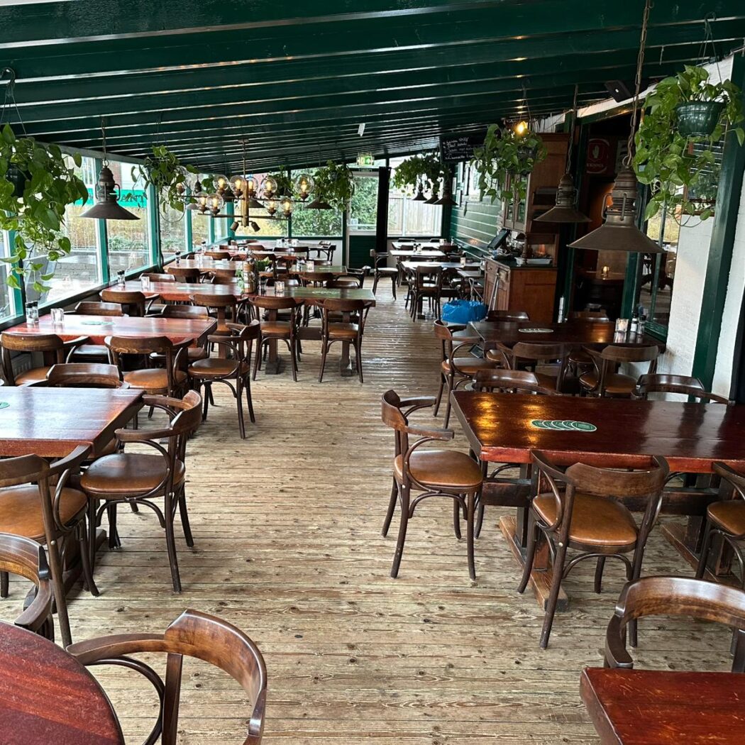 Leeg café-restaurant met rustieke houten tafels, stoelen, houten vloer en groene balken met planten.