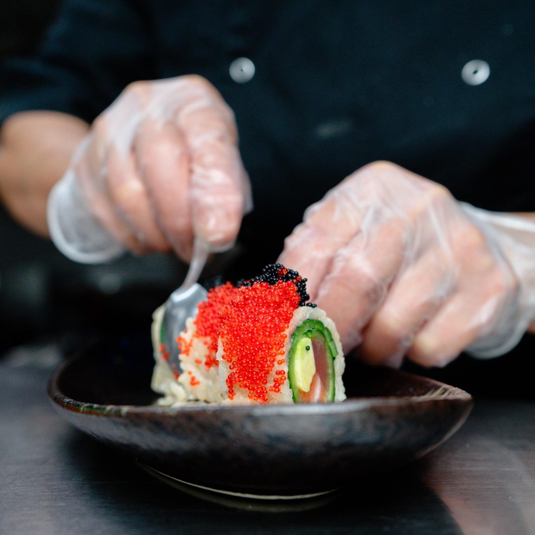Een chef met handschoenen decoreert een sushirol met rode en zwarte viskuit op een donker bord.