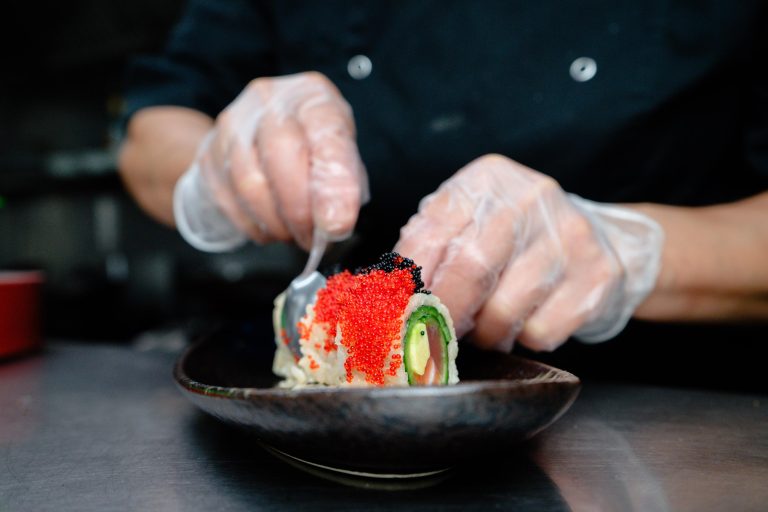 Een chef met handschoenen decoreert een sushirol met rode en zwarte viskuit op een donker bord.