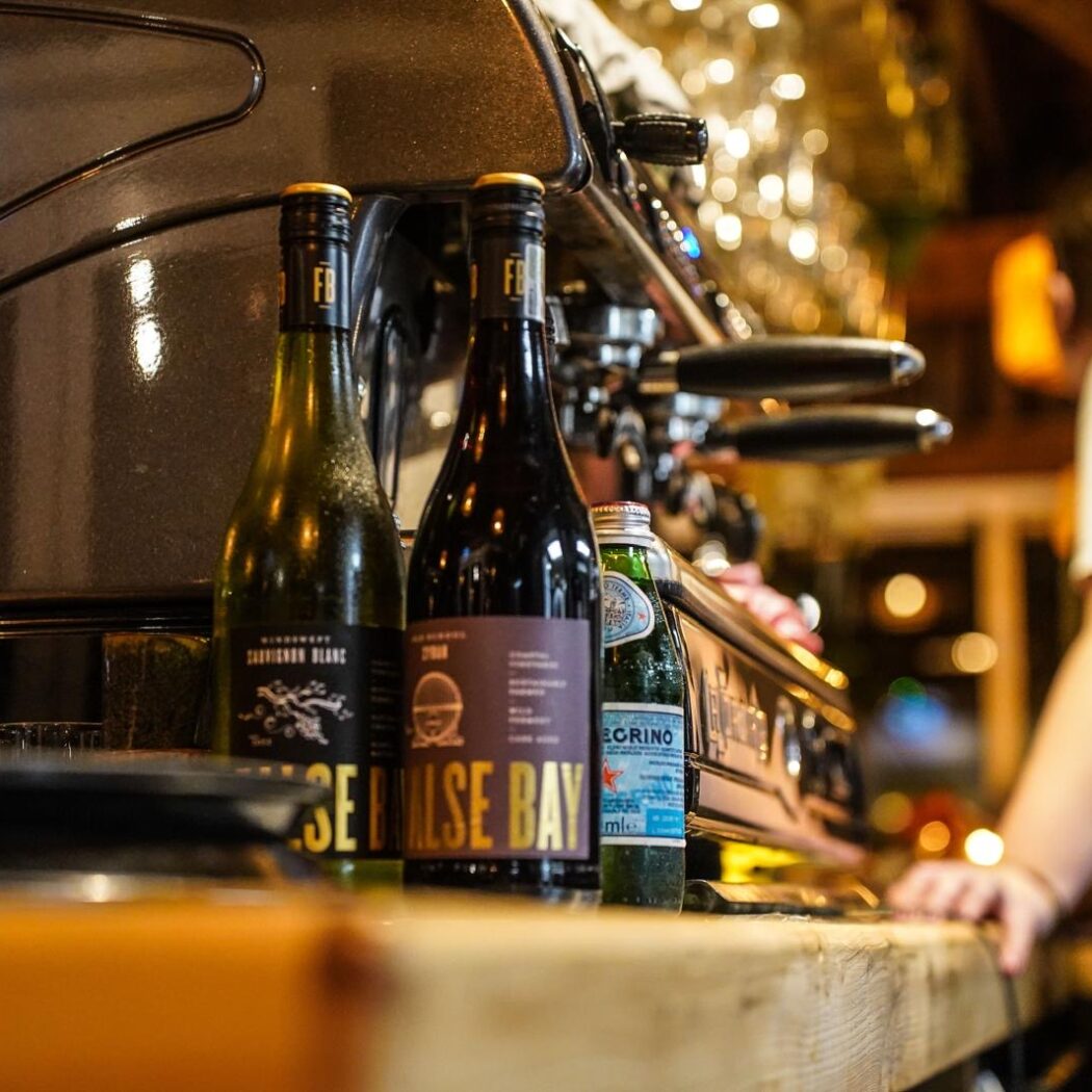 Twee False Bay wijnflessen en een S.Pellegrino fles staan op een houten bar naast een espressoapparaat, met een medewerker.