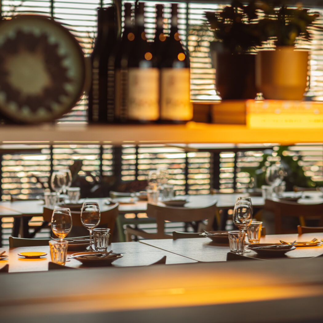 Sfeervol restaurantinterieur met gedekte houten tafels, wijnflessen en decor op een bovenliggende plank.